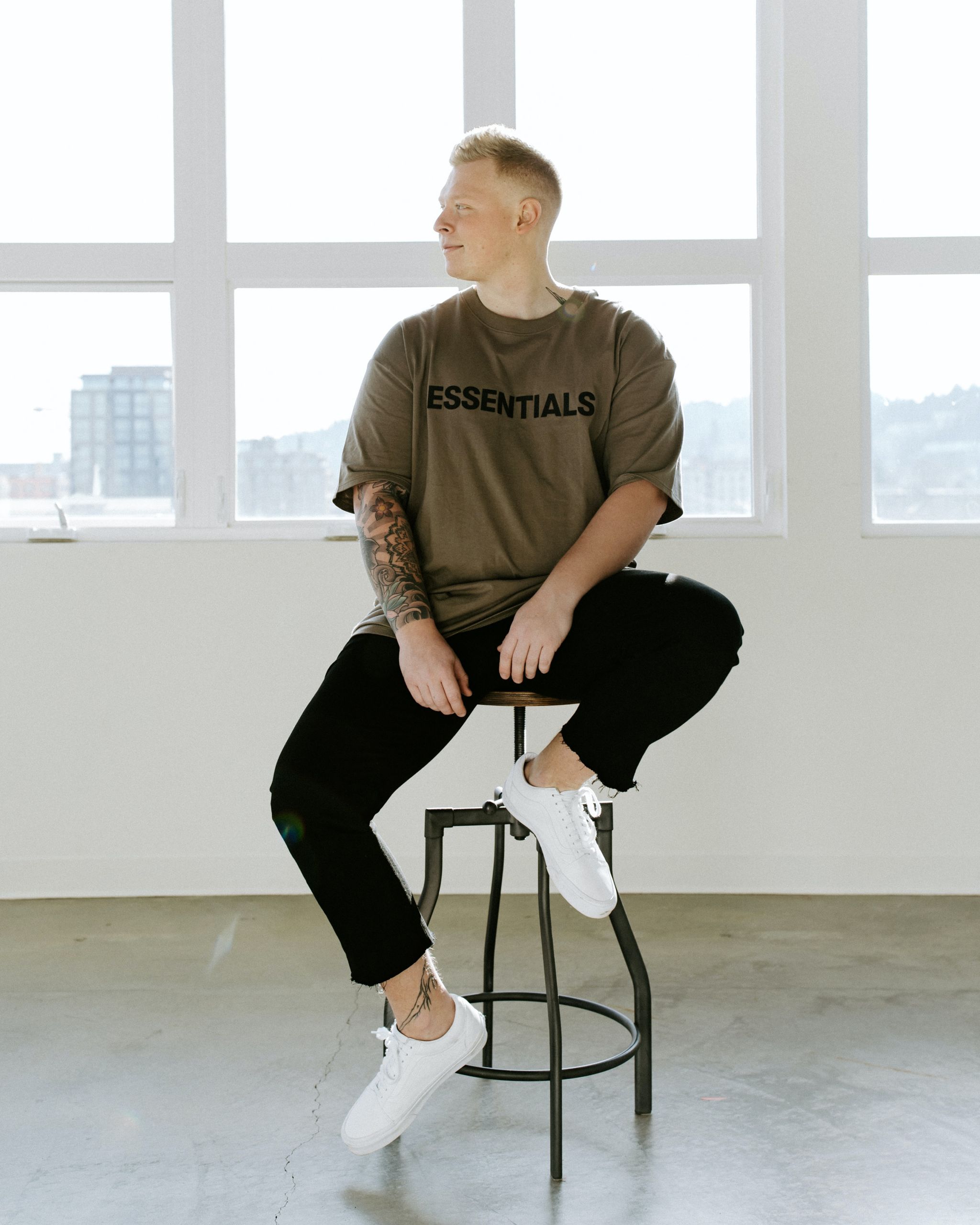 Eine Person mit kurzen Haaren sitzt auf einem hohen Hocker vor großen Fenstern. Sie trägt ein braunes „Essentials“-T-Shirt, schwarze Hosen und weiße Turnschuhe. Auf ihrem rechten Arm ist ein Tattoo zu sehen. Der Hintergrund ist hell und minimalistisch.