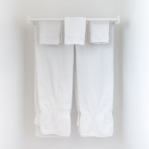 Ein Satz weißer Handtücher hängt ordentlich an einem Handtuchhalter vor einer schlichten weißen Wand. Zwei große Handtücher hängen nebeneinander, darüber liegen drei kleinere gefaltete Handtücher. Das Gesamtbild ist sauber und ordentlich.