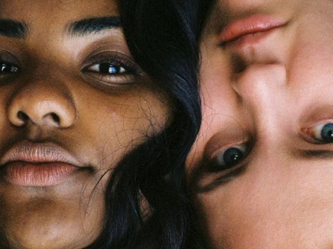 Nahaufnahme von zwei Personen mit unterschiedlichem Hautton, die ihre Köpfe dicht aneinander legen. Eine Person ist aufrecht und die andere kopfüber. Beide haben einen neutralen Ausdruck, der die Details ihrer Gesichter einfängt.
