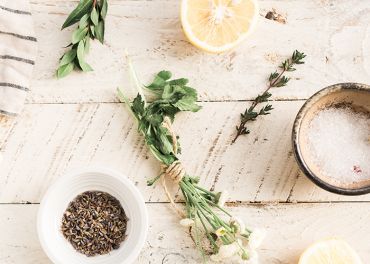 Ein Holztisch mit einer halbierten Zitrone, einer kleinen Schale mit Salz, einem Bündel frischer Kräuter, die mit Bindfaden zusammengebunden sind, einer Schale mit getrocknetem Lavendel und verstreuten Lorbeerblättern. Links ist teilweise ein gestreiftes Tuch sichtbar.