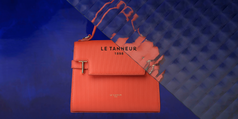 Eine stilvolle orangefarbene Le Tanneur-Handtasche steht vor einem leuchtend blauen strukturierten Hintergrund. Die Tasche hat ein elegantes Design mit Klappverschluss und dem eingeprägten Markennamen „Le Tanneur 1898“.