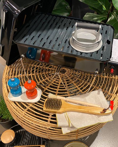 Ein Grill mit einem Brenner auf einem Korbtisch, dazu eine Grillbürste aus Holz, ein Tuch und kleine blaue und orangefarbene Gewürzmühlen auf einem Tablett. Im Hintergrund sind Pflanzen zu sehen.