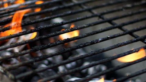 Nahaufnahme eines Grills mit heißen Kohlen darunter und kleinen Flammen, die zwischen den Metallrosten flackern. Der Grill weist einige verkohlte Überreste auf, was darauf hindeutet, dass er vor kurzem zum Kochen verwendet wurde.