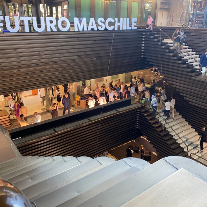 Ein mehrstöckiger Innenraum mit Holztreppen und Menschen, die den Raum erkunden. Auf einem großen Schild steht „Futuro Maschile“. Durch Glasfenster in den oberen und unteren Stockwerken sind verschiedene Ausstellungen und Aktivitäten sichtbar.
