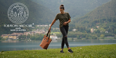 Eine Person steht auf einer Wiese an einem See und hält eine braune Einkaufstasche in der Hand. Sie trägt ein grünes T-Shirt und zerrissene Jeans. Im Hintergrund sind Berge und Häuser zu sehen. Links ist ein Logo mit „For People Who Care“ und „Made in Austria“ zu sehen.