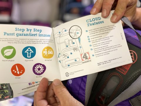 Eine Person hält eine aufgeschlagene Broschüre mit bunten Symbolen für Nachhaltigkeit, Ergonomie, Sicherheit, Design und Qualität in der Hand. Auf der rechten Seite sind die „CLOUD-Features“ mit einem Diagramm detailliert beschrieben. Im Hintergrund ist ein Rucksack zu sehen.