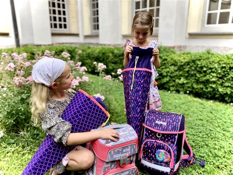 Zwei junge Mädchen sitzen auf einer Wiese in einem Garten und inspizieren ihre bunten Rucksäcke. Eine trägt ein Stirnband und hält eine lila Isomatte in der Hand, die andere schaut in ihren rosa-lila Rucksack. Im Hintergrund sind Blumenbüsche und Fenster zu sehen.