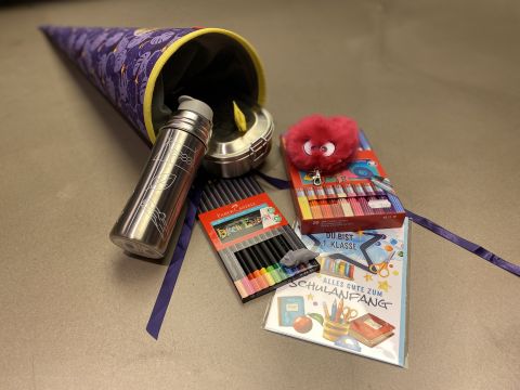 Ein großer, mit einem violetten Muster verzierter Kegel ist mit Schulsachen gefüllt, darunter Buntstifte, eine Metallwasserflasche, ein rotes Plüschtier und Grußkarten. Der Kegel und die Sachen sind auf einer flachen Oberfläche angeordnet.