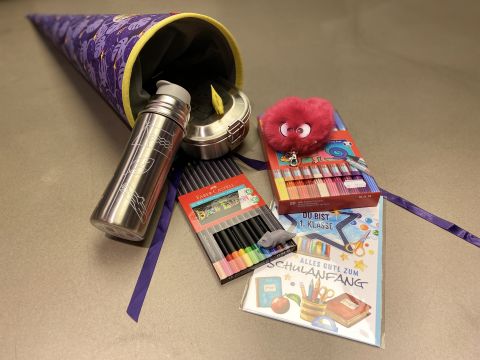 Eine große, bunte Schultüte ist geöffnet und gibt den Blick frei auf den Inhalt, wie eine Metalltrinkflasche, Buntstifte, ein rosa Plüschtier und ein Buch mit deutschem Text. Die Tüte ist mit Sternen verziert und mit einem lila Stoffbezug versehen.