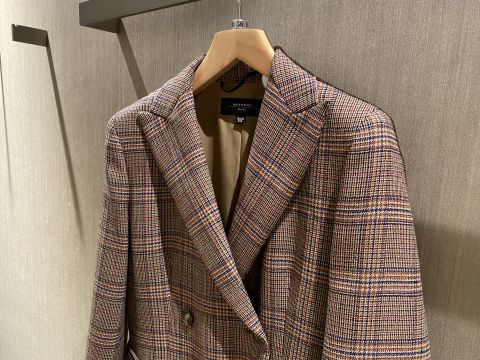 Ein karierter Blazer in Braun und Beige hängt auf einem hölzernen Kleiderbügel vor einer strukturierten beigen Wand. Der Blazer hat ein klassisches Karomuster mit Knopfverschluss und eine maßgeschneiderte Passform.