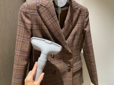 Eine Hand hält einen Dampfglätter und drückt ihn gegen einen karierten Zweireiher-Blazer, der an einem grauen Kleiderbügel hängt. Der Blazer weist ein klassisches Karomuster in Brauntönen auf. Die Szene spielt in einem Innenbereich.