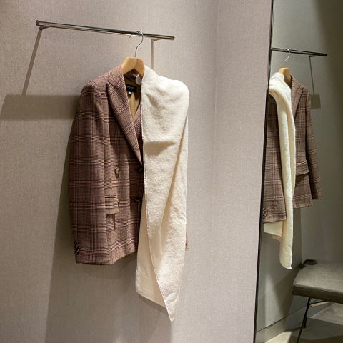 Ein braun karierter Blazer mit einem cremefarbenen Schal hängt an einem Ständer in einer Umkleidekabine. Ihr Spiegelbild ist im Spiegel zu sehen, und in der Ecke steht ein Hocker.