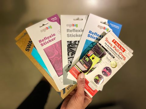 Eine Hand hält fünf Packungen Ergobag-Reflex- und Neonaufkleber in verschiedenen Farben und Mustern, darunter rosa, blaue und mehrfarbige Designs. Der Hintergrund ist eine verschwommene Innenkulisse.