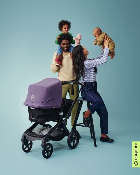 Eine fröhliche Familienszene mit einem Mann, der ein Kind auf seinen Schultern trägt, und einer Frau, die ein weiteres Baby hochhebt. In der Nähe steht ein lila Kinderwagen vor einem blaugrünen Hintergrund. Unten rechts ist das Bugaboo-Logo zu sehen.