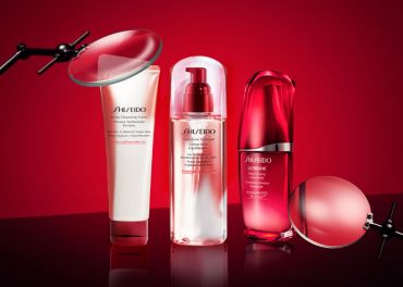 Drei Shiseido-Hautpflegeprodukte werden vor einem roten Hintergrund präsentiert, begleitet von einer Lupe, die die Details der Flaschen hervorhebt. Die Produkte umfassen einen Reinigungsschaum, einen Weichmacher und ein Konzentrat, jeweils in einer eleganten rot-weißen Verpackung.