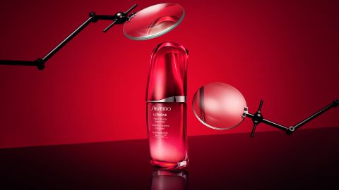 Eine rot-silberne Flasche mit einem Hautpflegeprodukt mit der Aufschrift „Shiseido“ steht zentriert vor einem leuchtend roten Hintergrund. Um die Flasche herum sind zwei schwarze Lupen aus Metall angebracht, die das elegante Design hervorheben.