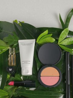 Verschiedene Make-up- und Hautpflegeprodukte, darunter eine Gesichtsmaske, Rouge, Lippenstift und Ampullen, angeordnet auf grünen tropischen Blättern.