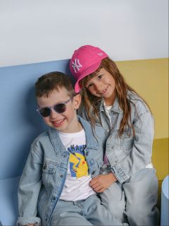 Zwei junge Kinder in Jeansjacken sitzen auf einer bunten Bank. Der Junge trägt eine Sonnenbrille und ein Pikachu-Shirt, während das Mädchen eine rosa Mütze trägt und lächelt.