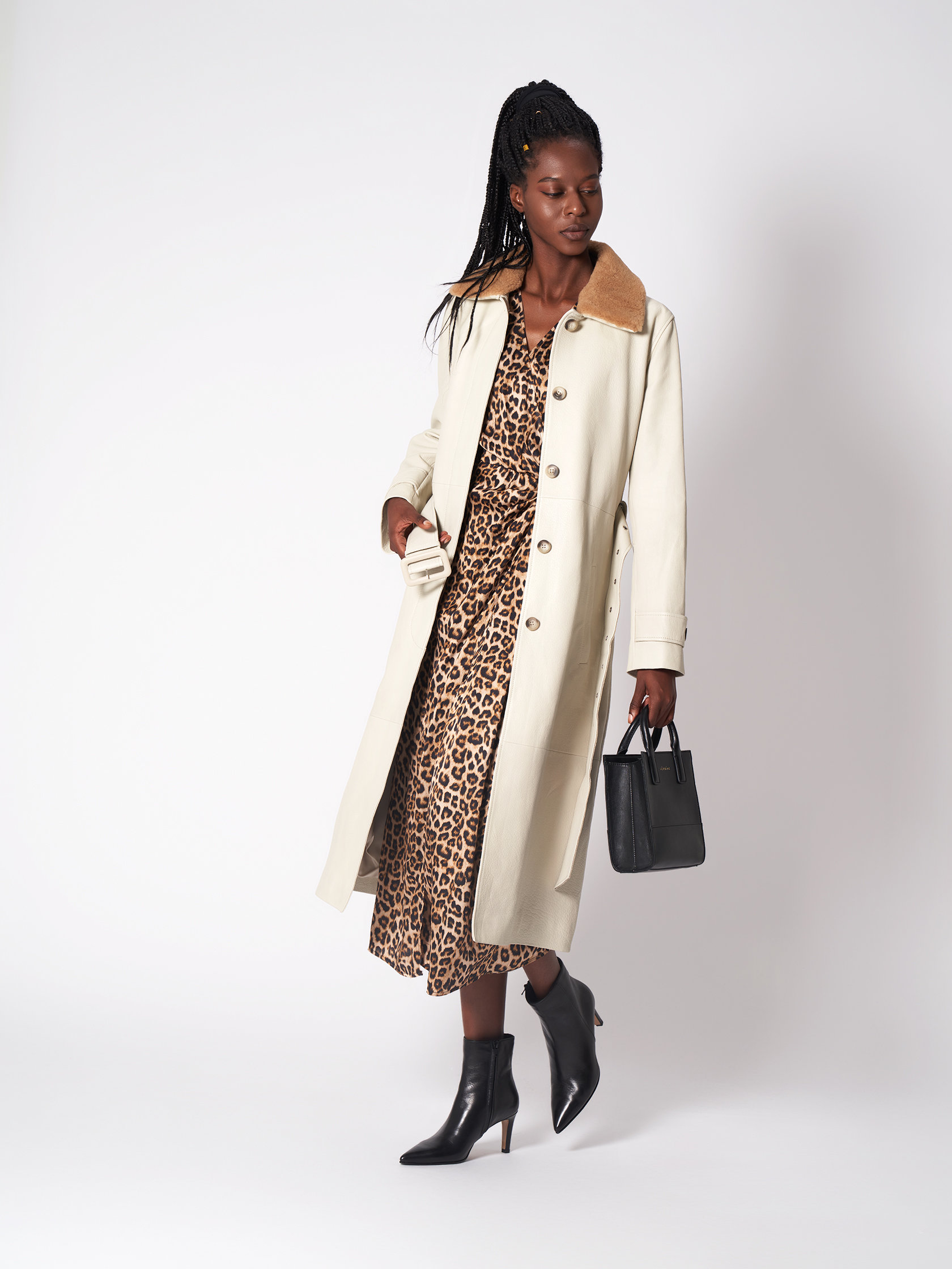 Eine Frau trägt einen langen beigen Mantel über einem Kleid mit Leopardenmuster und hält eine kleine schwarze Handtasche. Ihr Haar ist zu langen Zöpfen geflochten und sie trägt schwarze Stiefeletten. Der Hintergrund ist schlicht weiß.