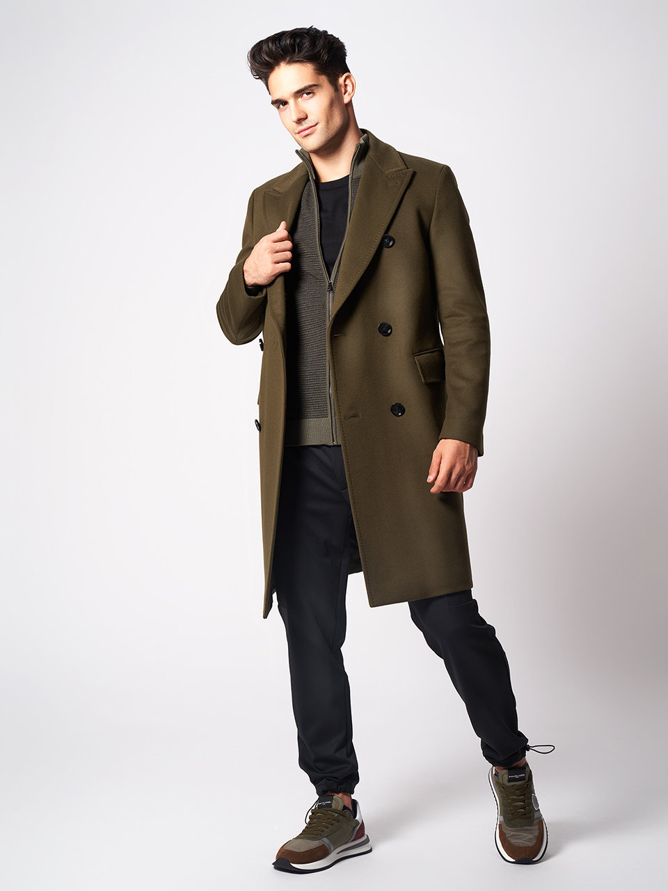 Ein Mann steht vor einem schlichten Hintergrund. Er trägt einen olivgrünen Mantel über einem dunklen Pullover und einer schwarzen Hose. Seine Hand liegt auf dem Revers des Mantels, ein Bein ist leicht angewinkelt. Er trägt lässige braun-weiße Turnschuhe.