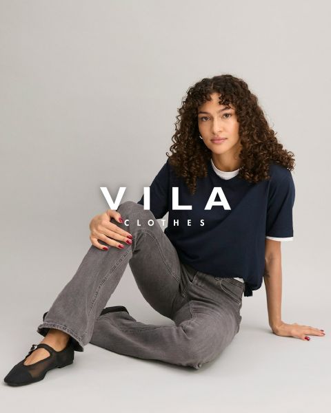Eine Person mit lockigem Haar sitzt vor einem neutralen Hintergrund auf dem Boden und trägt ein marineblaues T-Shirt, graue Jeans und schwarze Schuhe. Der Text „VILA CLOTHES“ ist über das Bild gelegt.