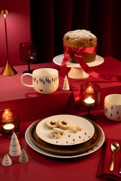 Eine festlich gedeckte Tafel mit einem großen Kuchen, der mit einer roten Schleife umwickelt und mit Puderzucker bestäubt ist, auf einem Ständer. Es gibt Kekse auf Tellern, Themenbecher, Kerzen, rot-weißen Christbaumschmuck und einen tiefroten Hintergrund.