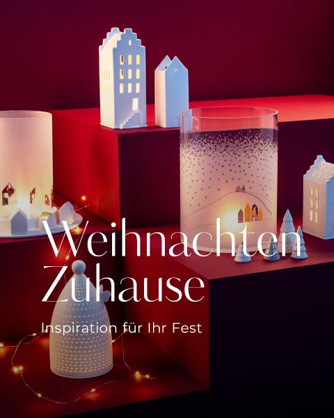 Eine festliche Auslage mit weißen Miniaturhäusern, Kerzenhaltern aus Glas und Lichterketten auf roten Podesten. Der Text lautet: "Weihnachten Zuhause. Inspiration für Ihr Fest." Die Szene strahlt eine warme, gemütliche Weihnachtsstimmung aus.