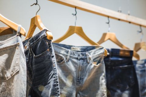 Eine Kollektion von Jeans in verschiedenen Farbtönen hängt auf hölzernen Kleiderbügeln an einer Holzstange. Die Jeans sind ordentlich angeordnet und zeigen eine Reihe von Stilen und Waschungen vor einem weichen, neutralen Hintergrund.