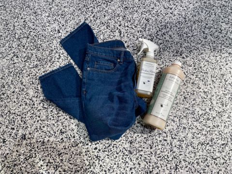 Eine Bluejeans liegt auf einem gesprenkelten grau-schwarzen Untergrund. Neben der Jeans liegen zwei Reinigungsprodukte: eine Sprühflasche und eine Flasche Waschmittel, beide mit minimalistischen Etiketten.