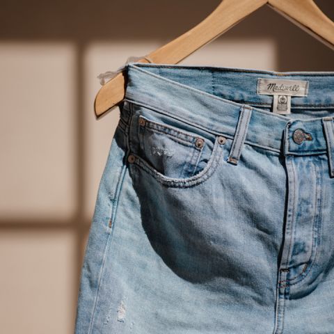 Nahaufnahme einer hellblauen Jeans, die an einem hölzernen Kleiderbügel hängt. Im Hintergrund ist ein Sonnenfleck zu sehen, der ein Gittermuster auf der beigen Wand erzeugt. Die Jeans weist in der Nähe der Taschen leichte Abnutzungsspuren auf.