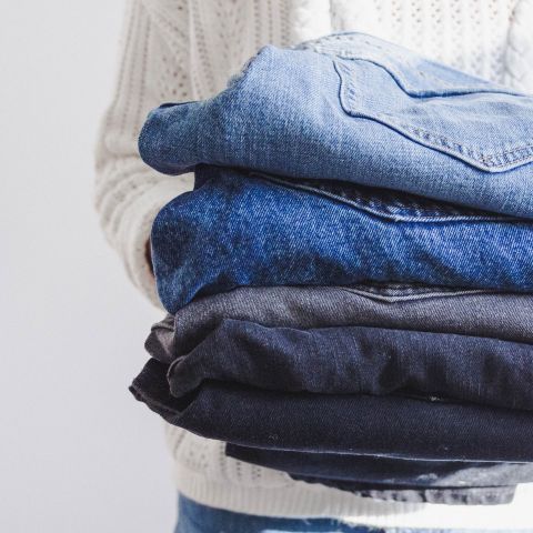 Eine Person hält einen Stapel gefalteter Jeans. Die Jeans haben verschiedene Blau- und Schwarztöne. Die Person trägt einen weißen Strickpullover mit sichtbarer Textur im Hintergrund.