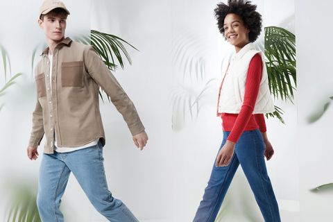 Ein Mann in beiger Jacke, blauen Jeans und beiger Mütze und eine Frau in weißer Weste, rotem Pullover und blauen Jeans gehen selbstbewusst in einer hellen, stilvollen Umgebung mit großen grünen Blättern im Hintergrund.