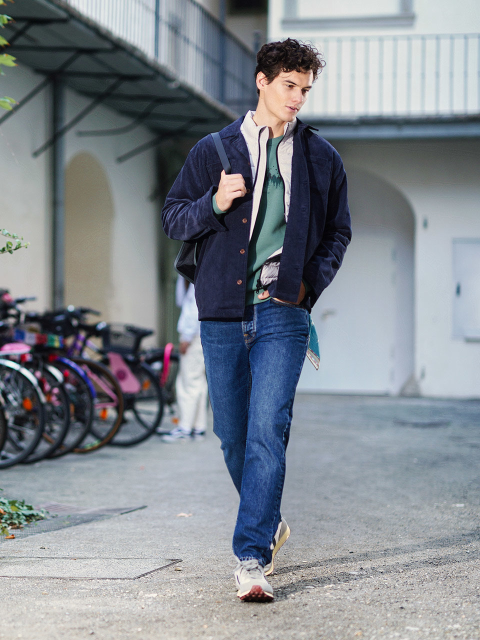 Eine Person läuft selbstbewusst auf einer Straße. Sie trägt eine marineblaue Jacke, einen grünen Pullover und blaue Jeans. Über der Schulter trägt sie eine schwarze Tasche. Im Hintergrund sind Fahrräder und ein Gebäude zu sehen.