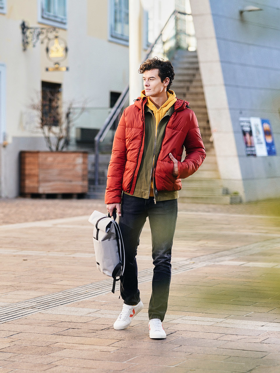 Eine Person in einer roten Pufferjacke und dunklen Jeans geht mit einem hellen Rucksack eine Straße entlang. Sie kommen an einem Gebäude vorbei, an dessen Wänden Plakate hängen. Die Umgebung wirkt urban und leger.