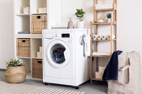 Eine aufgeräumte Waschküche mit einer Frontlader-Waschmaschine. Auf den Regalen stehen geflochtene Körbe, cremefarbene Handtücher und Topfpflanzen. Auf einem Holzregal sind Toilettenpapier und Seife ausgestellt. Daneben steht ein Wäschekorb mit einem blauen Handtuch. Auf dem Boden liegt ein gemusterter Teppich.