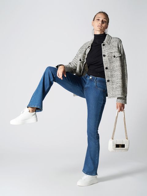 Eine Person posiert in einem trendigen Outfit. Sie trägt einen schwarzen Rollkragenpullover, eine karierte Jacke, blaue Jeans und weiße Turnschuhe. Sie hält eine weiße Handtasche und hat ein Bein hochgehoben. Sie steht vor einem schlichten Hintergrund.
