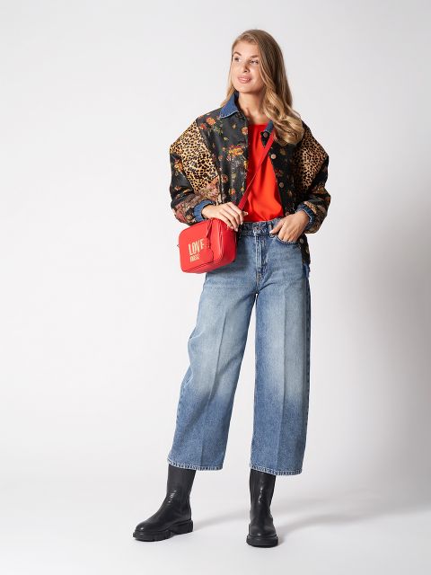 Eine Person trägt eine Jacke mit Blumen- und Leopardenmuster über einem roten Oberteil, blaue Jeans mit weitem Bein und schwarze Stiefel. Sie trägt eine rote Umhängetasche mit der Aufschrift „LOVE“ in weißer Schrift auf einem schlichten weißen Hintergrund.