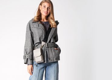 Eine Person trägt eine schicke graue Jacke und blaue Jeans und hält eine beige Umhängetasche. Der Hintergrund ist schlicht und neutral und unterstreicht das modische Outfit.