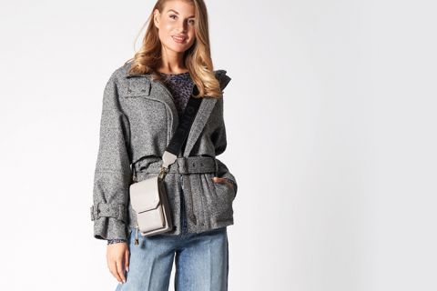 Eine Person trägt eine schicke graue Jacke und blaue Jeans und hält eine beige Umhängetasche. Der Hintergrund ist schlicht und neutral und unterstreicht das modische Outfit.