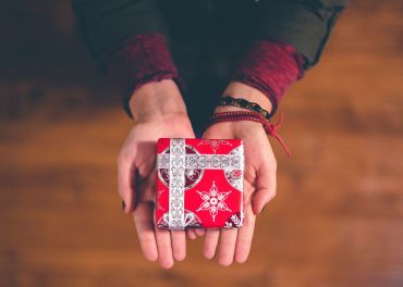 Eine Person mit roten Ärmeln hält eine kleine rote Geschenkbox mit silbernem festlichem Muster und weißer Schleife und bietet sie nach vorne an. Der Hintergrund ist unscharf, wodurch das Geschenk in den Händen der Person hervorgehoben wird.