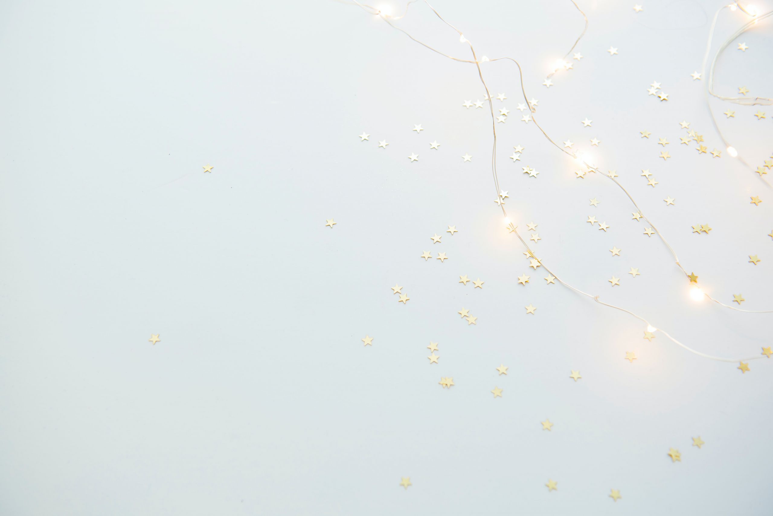 Ein weicher, hellblauer Hintergrund mit verstreutem, kleinem, goldenem Konfetti in Sternform. Zarte, leuchtende Lichterketten sind ineinander verschlungen und schaffen eine verträumte, festliche Atmosphäre.
