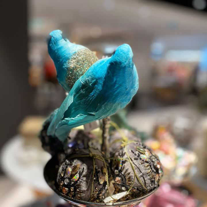 Zwei dekorative blaue Vögel sitzen auf einem mit Strasssteinen und Glitzer verzierten Ast. Der Hintergrund ist sanft verschwommen, wodurch die leuchtenden Farben und komplizierten Details der Vögel hervorgehoben werden.