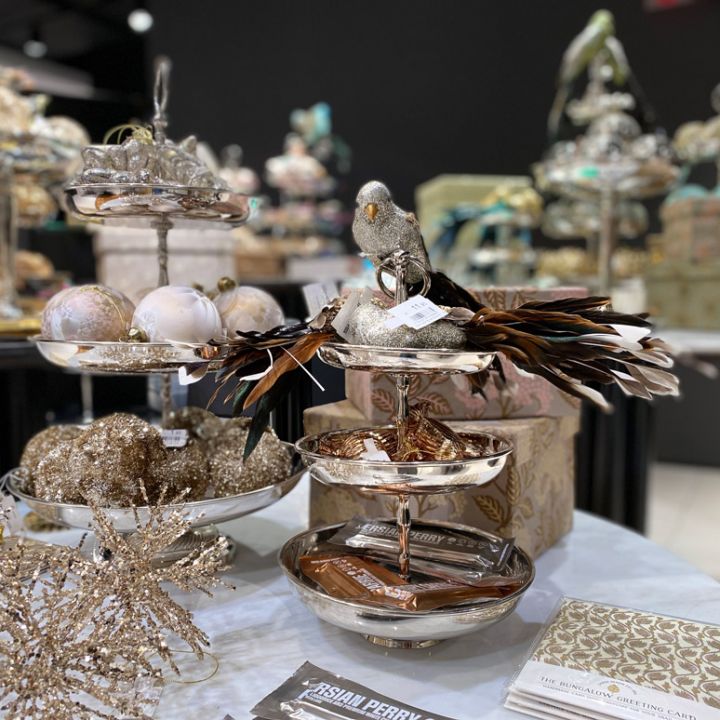 Eine Ausstellung von Weihnachtsdekorationen mit mehrstöckigen silbernen Tabletts, die mit glitzernden Ornamenten geschmückt sind, einer Vogelfigur mit Federn und dekorativen Objekten. Im Hintergrund sind weitere festliche Tischdekorationen zu sehen.