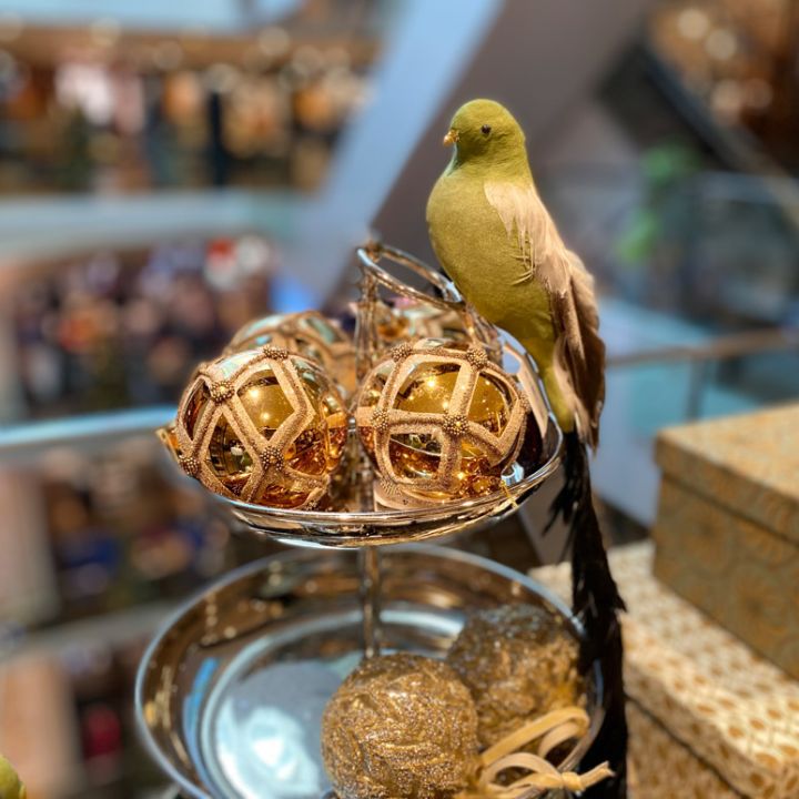 Ein dekorativer grüner Vogel thront auf einem Tablett mit goldenen, glitzernden Ornamenten in einer Ladenauslage. Der Hintergrund zeigt eine verschwommene Ansicht eines mehrstöckigen Einkaufszentrums.