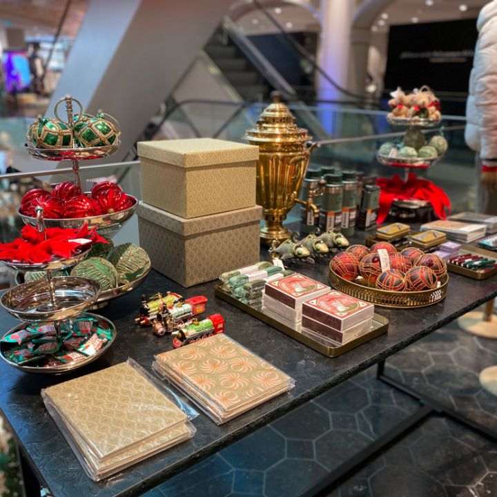 Auf einem Ausstellungstisch in einem Geschäft sind verschiedene Weihnachtsdekorationen und Geschenke zu sehen, darunter Weihnachtsschmuck, verpackte Schachteln und Süßwaren. Im Hintergrund sind eine Rolltreppe und Schaufensterpuppen in Winterkleidung zu sehen.