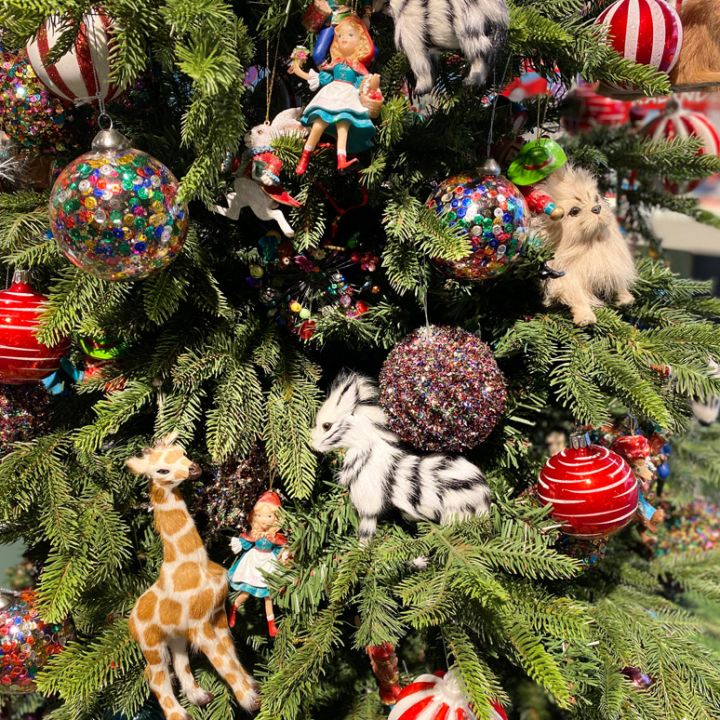 Ein Weihnachtsbaum, der mit farbenfrohen Ornamenten geschmückt ist, darunter gemusterte Kugeln, Stofftiere wie eine Giraffe und ein Zebra sowie kleine Puppen. Der Baum ist üppig mit grünen Zweigen und farbenfrohen Dekorationen geschmückt und schafft eine festliche Weihnachtsszene.