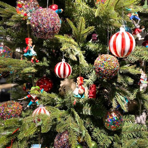 Ein festlicher Weihnachtsbaum, geschmückt mit bunten Kugeln, gestreiften Ornamenten in Zuckerstangenoptik und kleinen Elfenfiguren. Eine neugierige Katze hat es sich zwischen den Zweigen gemütlich gemacht und ist teilweise von den Dekorationen verdeckt.