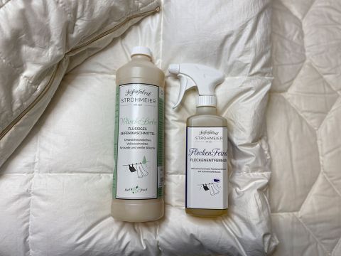 Auf einer weißen gesteppten Oberfläche stehen zwei Flaschen mit Reinigungsmitteln. Eine ist ein Flüssigwaschmittel in einer hohen Flasche, die andere ein Fleckenentferner-Spray in einer kleineren Flasche. Beide haben weiße Etiketten mit deutschem Text.