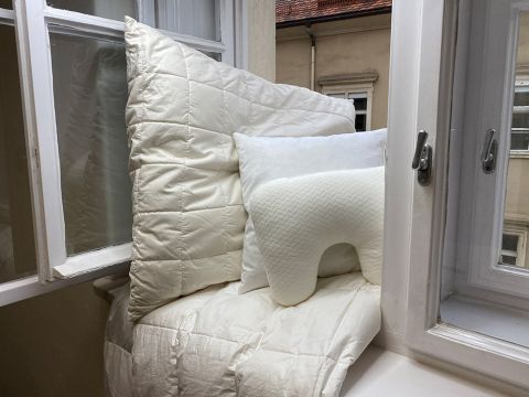 Gemütliche Fensterecke mit flauschigen weißen Kissen und einer Steppdecke auf einem Sims. Zwei offene Fenster geben den Blick auf ein Gebäude von außen frei. Zu den Kissen gehören ein großes quadratisches, ein kleineres rechteckiges und ein U-förmiges Reisekissen.