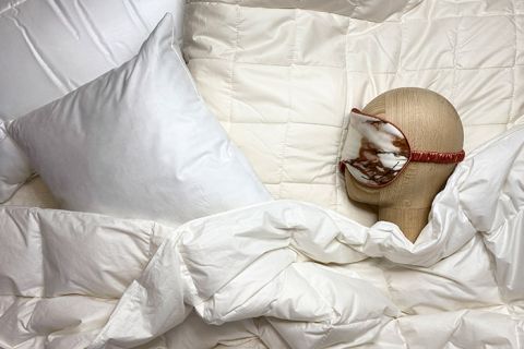 Ein Puppenkopf mit Schlafmaske ruht auf einem Bett, das mit einer weißen Bettdecke und einem Kissen bedeckt ist. Die Umgebung vermittelt eine gemütliche und erholsame Atmosphäre.